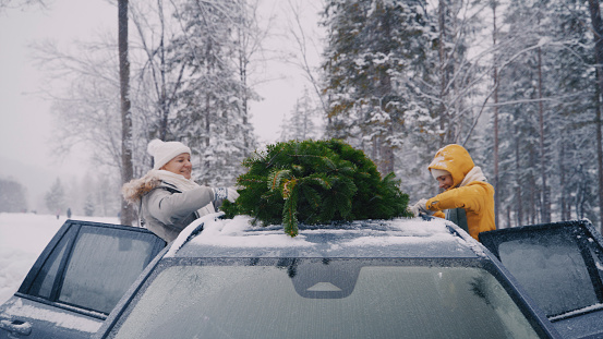 Two women friends tying christmas tree on top of car roof on snowy forest landscape during winter