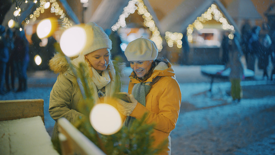Two happy women friends in warm clothing using mobile phone at snowy christmas market