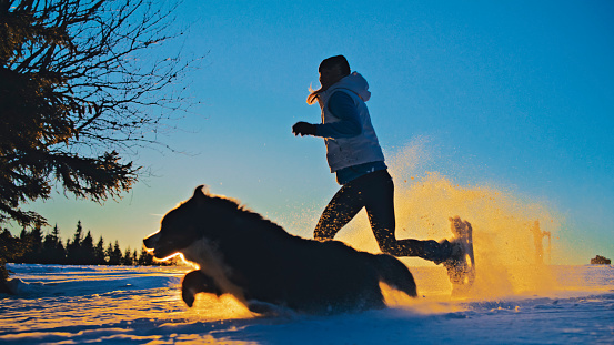 Carefree woman in warm clothing snowshoeing with her pet dog on snowy slope during dawn