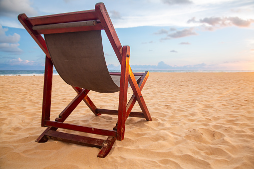 A wooden beach chair on a sandy beach in sunset. The chair is made of wood and has a brown canvas seat. The chair is facing the ocean and the horizon. The background consists of the ocean, the horizon, and a cloudy sky. The sand is light in color and has small ripples in it.