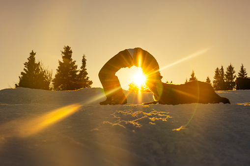 Silhouette of woman practicing bridge pose on snowy hill with her pet Bernese mountain dog during sunset