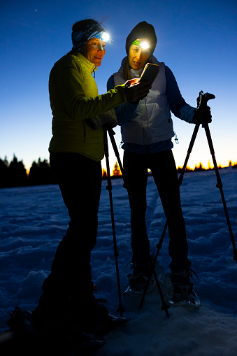 Two women skiers with flashlight on forehead and ski poles in hands using mobile phone in snow during night