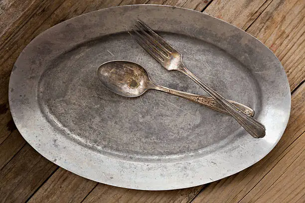 An overhead close up shot of an old pewter platter and some tarnished silverware. Shot on a grungy old wooden table.