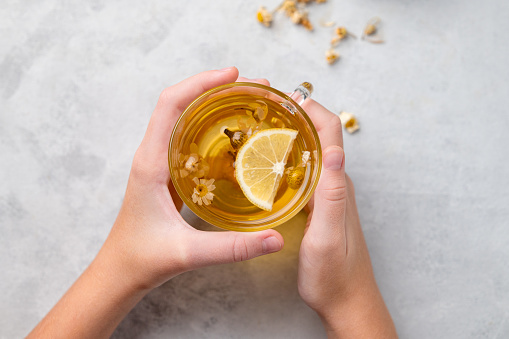 Hands holding a glass cup with chamomile herbal tea on a light background. The concept of a healthy drink for health and immunity. Top view and copy space.