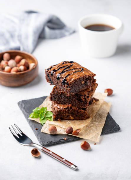 A stack of chocolate brownies with mint, hazelnuts and salted caramel on baking paper on a light background with a cup of coffee and nuts.