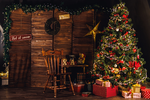 Wide shot of a cozy, rustic Christmas set up with a glistening Christmas tree, stacked gifts and empty chair and lots of decorations.