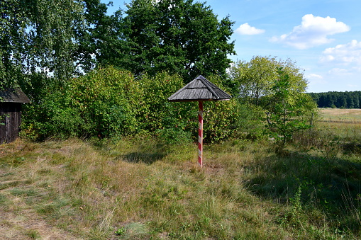 A close up on a stand with a tiled roof painted red and white standing in the middle of a small grove or forest and being overgrown with trees spotted next to a vast field on a sunny summer day