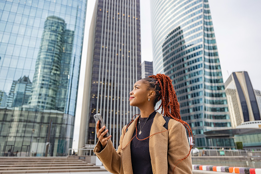 Waist up side shot from a low angle with blurred background of a young and beautiful African woman walking through the financial district of the city while holding her phone to find the address of her upcoming job interview.