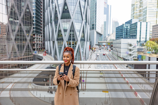 Aerial waist up front shot with blurred background of an African woman standing using her mobile phone on a bridge overlooking the financial district on a cold morning in Paris.