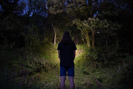 Back view of young man wearing shorts and t-shirt and illuminating forest with head lamp on summertime
