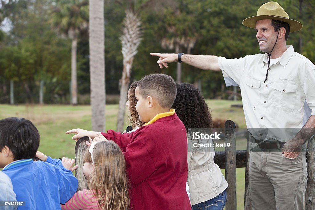 Zoo keeper with group of children at animal exhibit Zoo keeper (30s) with multi-ethnic group of elementary school children at zoo, standing on observation deck overlooking animal exhibit. Park Ranger Stock Photo