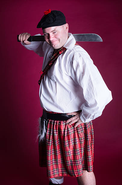 Smiling Scot with machete over shoulder. Vertical studio shot on burgundy of man in kilt with hand on hip. tam o'shanter stock pictures, royalty-free photos & images