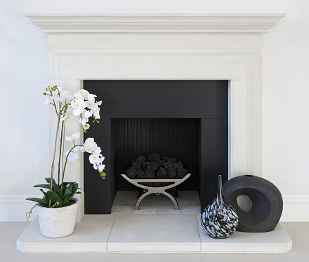 an elegant white gas fireplace in a luxury home with black marble surround and white marble base. The frame is in white painted wood. A silk white orchid sits to the left whilst two decorative vases are on the right. The fireplace itself is a stainless steel cradle-type with mock coals.