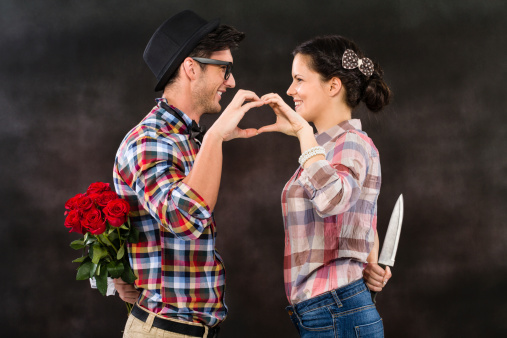 Retro young couple making heart shape with their hands. Man holding bouquet of red roses behind his back while his girlfriend is holding table knife behind her back.