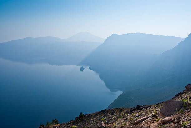 Early Morning Fog Over Crater Lake A combination of early morning fog and smoke from forest fires gives this scene a strange bluish glow. This photograph was captured from Garfield Peak in Crater Lake National Park, Oregon, USA. Phantom Ship and Mount Scott are in the distance. jeff goulden crater lake national park stock pictures, royalty-free photos & images