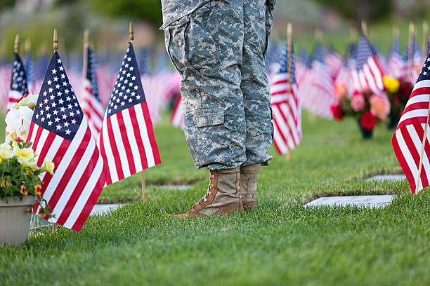 A soldier in uniform standing in solidarity with fallen soldiers at a cemetery with American flags beside each stone to commemorate Memorial Day.  Yellow flowers are close to the soldier's feet, and another set of colorful flowers are noticeable in the background.  The gravestones and flags are set in freshly cut green grass.