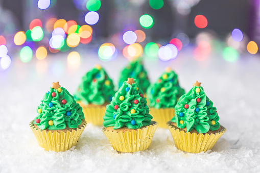 Cute tasty Christmas tree Christmas cupcakes, with gold paper cakes, on white snow with lights bokeh in the background