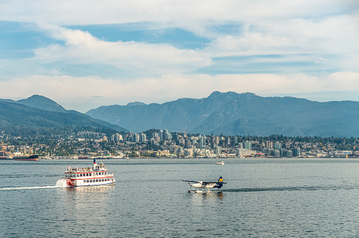 Vancouver, Canada - 10th September 2023: A paddleboat takes  tourists on a tour of Vancouver Harbour and Burrard Inlet while a Harbour Air seaplane comes in to land. View looks over the bay towards North Vancouver and Grouse Mountain.