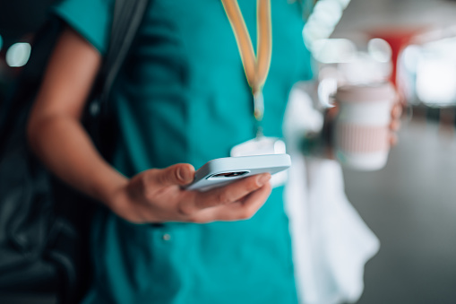 Close-up of a young Caucasian female nurse`s hands carrying a coffee cup and using her mobile phone. She is in scrubs and has her medical ID card on her, while carrying her lab coat and a backpack on her shoulder.