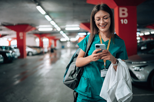 Young Caucasian female nurse standing in a parking lot in her scrubs, while typing on a mobile phone and cheerfully smiling.