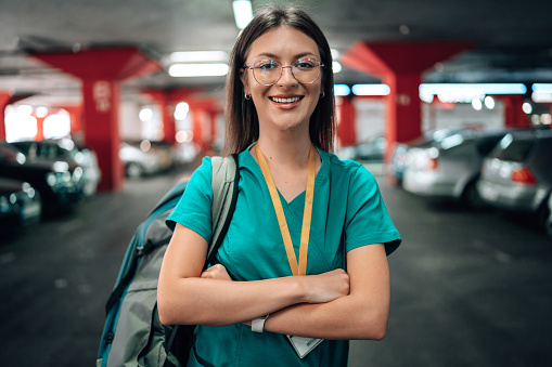 Portrait of a young Caucasian female nurse standing in a parking lot in her scrubs and cheerfully smiling, while looking at the camera. She has her hands crossed, while carrying a backpack on her shoulder.