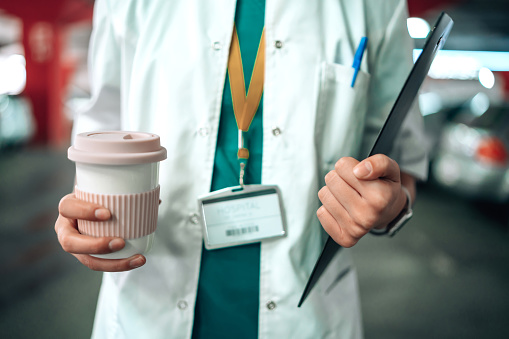 Close-up of a young Caucasian female nurse`s hands carrying a coffee cup and a note pad. She is standing in a parking lot in scrubs and a lab coat with her medical ID card around her neck.