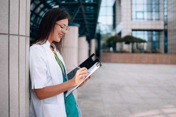 junge krankenschwester von der taille aufwärts steht in ihrem laborkittel und schrubbt sich an der wand eines krankenhausgebäudes, während sie fröhlich lächelt und auf ihren notizblock schreibt - healthcare and medicine smiling group of people lab coat stock-fotos und bilder