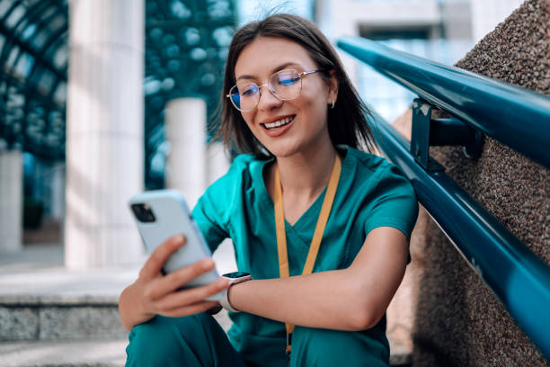 istock Young female nurse sitting on stairs in her scrubs, while cheerfully smiling and using her phone 1748387989