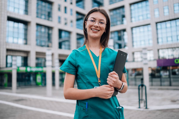 istock Young female nurse standing outside in scrubs, while holding a note pad and cheerfully looking at the camera 1748388554