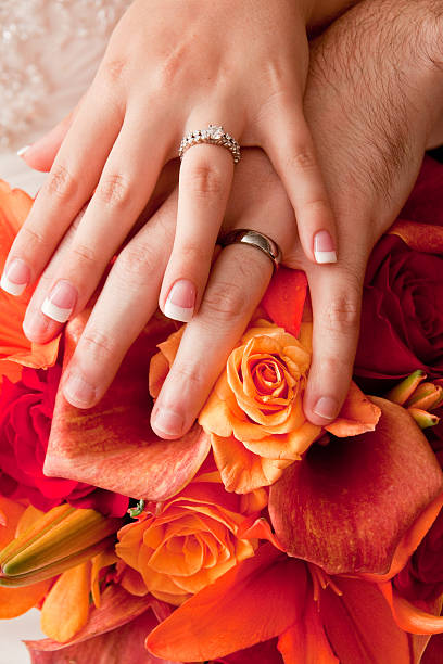 A bride and groom holding hands after the wedding.  They are wearing their wedding rings as a symbol of their love.  Their hands are resting on a beautiful bouquet of autumn colored roses and calla lilies.