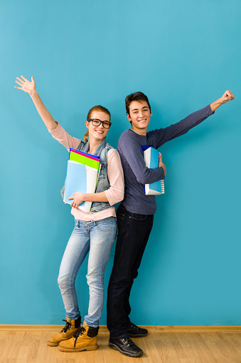 Teenage boy and teenage girl (age 15) holding books, standing with hands raised in front of the blue wall.