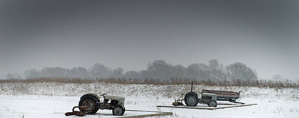 2 old tractors in a field with snow falling from leaden overcast skies.