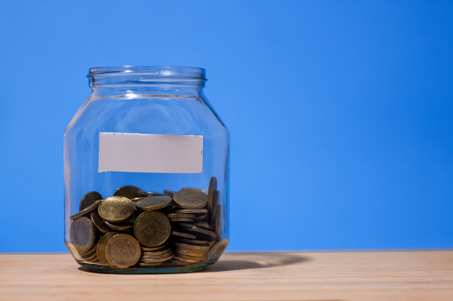 Glass jar full of coins with blank white label on it isolated on blue background with copy space illustrating savings for paying taxes or for vacation, pension funds, healthcare or house expenses.