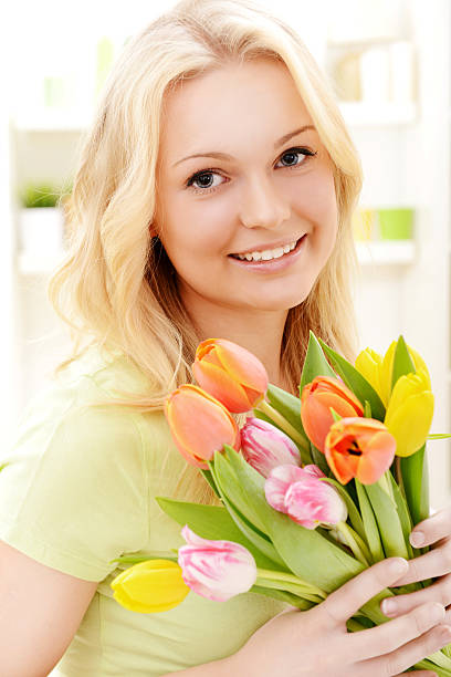 Portrait of a young blond hair woman wearing a blue turtleneck shirt standing in a studio with a blue background, playfully cowering her one eye with a yellow tulip flower and looking at camera with the other