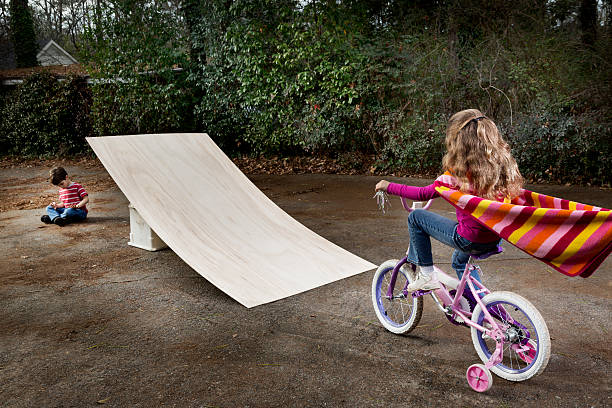 It Seemed Like a Good Idea "A little girl is about to jump a ramp on her bike, a small boy sits unaware on the far side." child behaving badly stock pictures, royalty-free photos & images