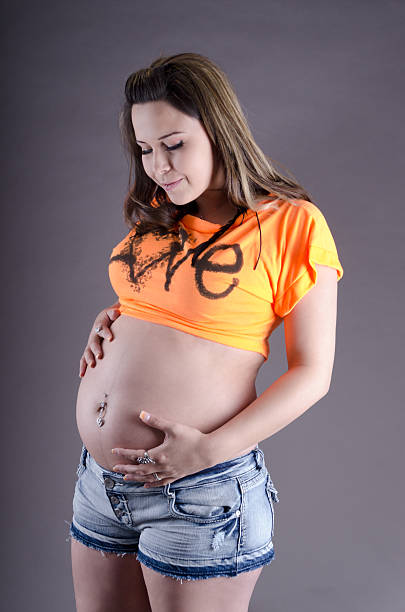 Young pregnant woman smiling down at belly. Vertical studio shot on gray of young woman in short t shirt and cutoffs holding belly. graphic t shirt stock pictures, royalty-free photos & images