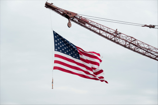 Conceptual image: Rebuilding America. A tattered American flag attached to a construction crane waves in the wind against a pale blue cloudy sky.