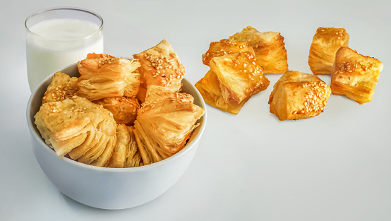 Bunch of freshly baked small square sesame puff pastry Zu-Zu, offered in a white ceramic bowl and tossed around, set with a glass of Yogurt on vignette white background, high resolution stock photo.