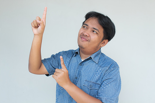 Young asian man in blue t-shirt with surprised and happy expression. While pointing above. Isolated on gray background.