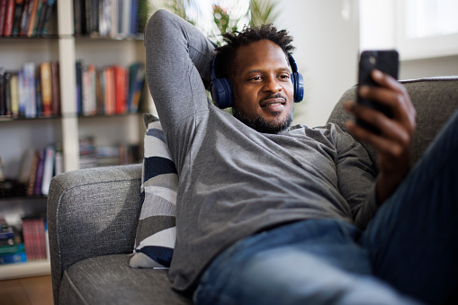 Smiling man with headphones resting on couch and using mobile phone