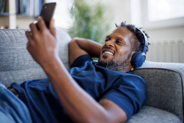 istock Smiling man with bluetooth headphones using mobile phone and relaxing on sofa 1748478741