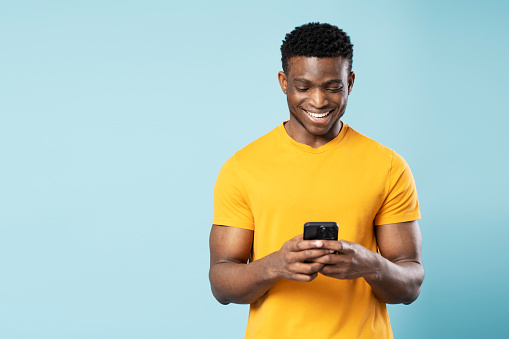 Handsome smiling African man holding smartphone using mobile app shopping online isolated on blue background. Technology concept