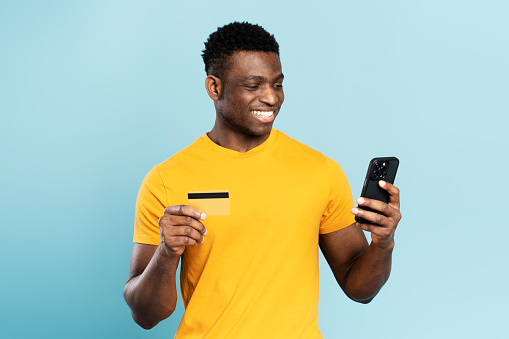 Portrait of handsome smiling African American man holding credit card, suing mobile app shopping online isolated on blue background. Happy stylish male ordering food. Technology concept