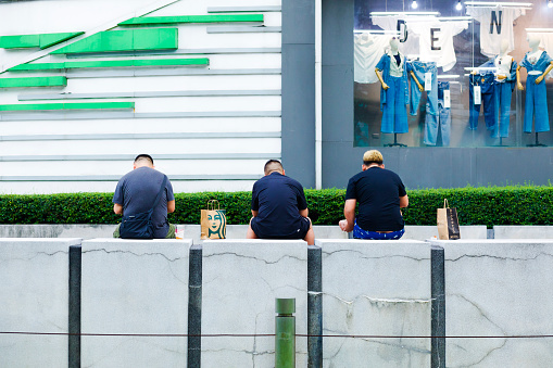 Three thai men with Starbucks bags  are sitting outside of Central Plaza  Rama 9 shopping mall at Ratchadaphisek Rd in Bangkok. Men have a break. They are sitting on a small wall. In background is facade of mall and a window of Robinson store.