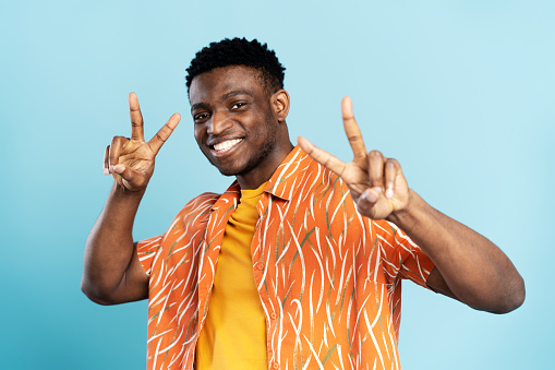 Portrait of handsome smiling African American man wearing orange shirt showing peace sign isolated on blue background. Young stylish hipster model looking at camera posing for pictures. Summer