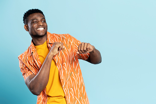 Handsome happy African American man wearing casual clothes dancing, having fun isolated on blue background. Positive lifestyle, summer concept