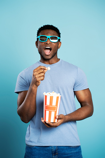 Excited nigerian man wearing 3D glasses eating popcorn watching movie isolated on blue background. Handsome hipster male in casual clothes holding paper box with snacks