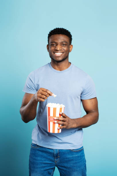 uomo africano positivo in maglietta blu che mangia popcorn isolato su sfondo blu, guardando la macchina fotografica - cardboard box immagine foto e immagini stock