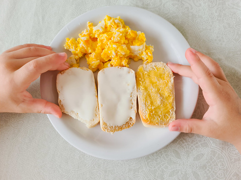 Child's hands having healthy breakfast. Scrambled eggs, bread with cream cheese and bread with butter.