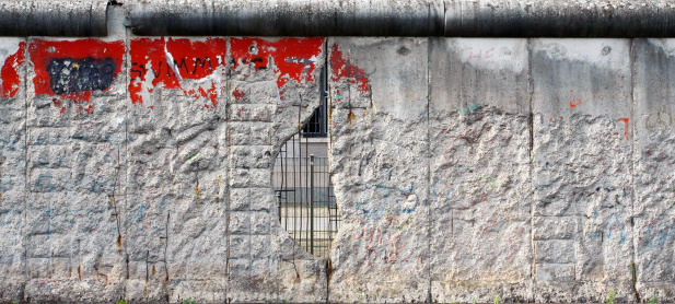 A hole in a remaining section of the Berlin Wall allows a view into what was formerly East Berlin.  The panorama format highlights the effect of segregation and the effect of the hole and its outward view.   This damaged section of wall has been retained in a Berlin outdoor museum.